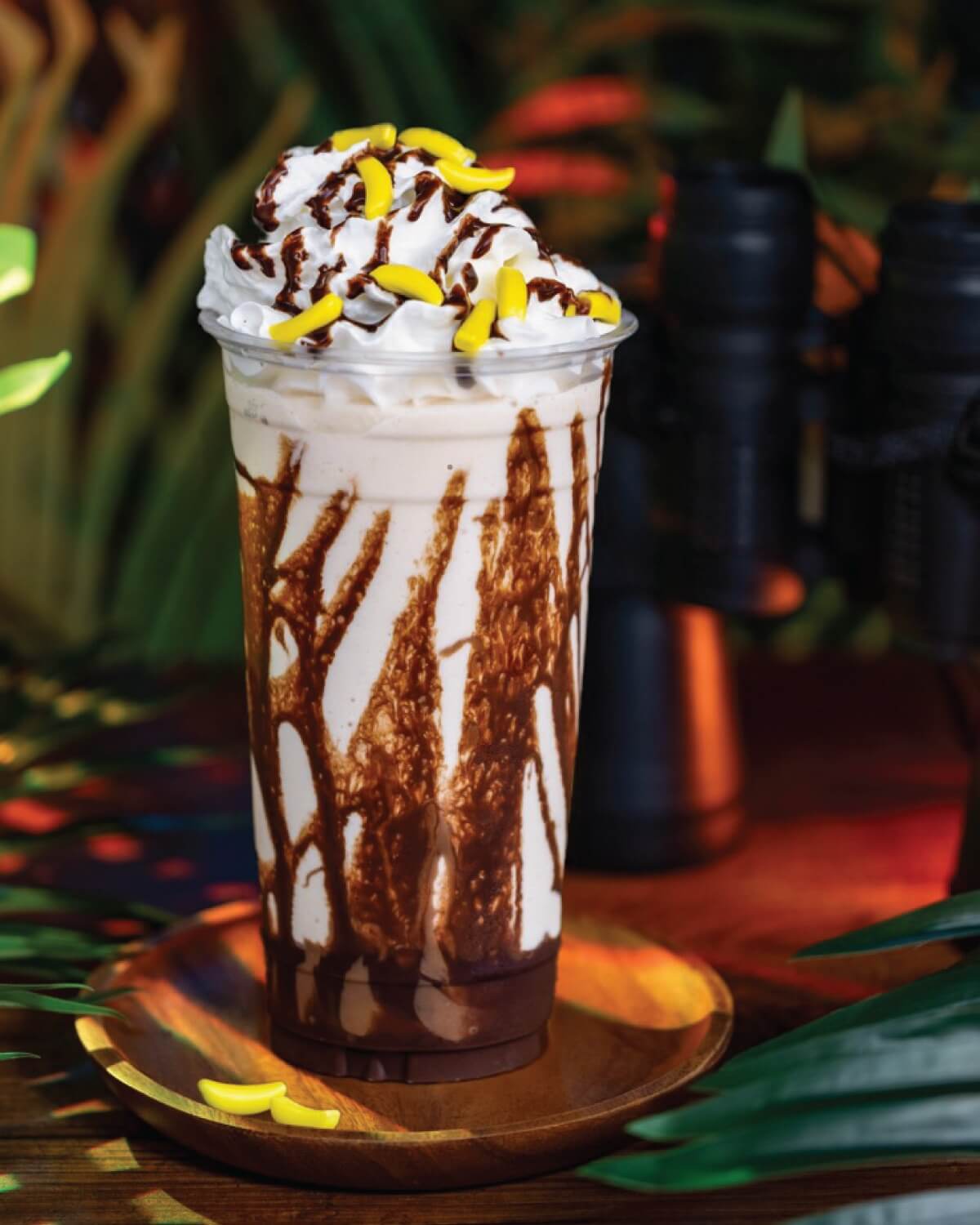 12 Florida Food & Heritage Trails Worth Traveling For - 8 A tall glass of iced chocolate drink topped with whipped cream, chocolate drizzle, and yellow candy pieces, placed on a wooden plate—perfect for travel breaks along Florida food heritage trails, with foliage and binoculars in the background.