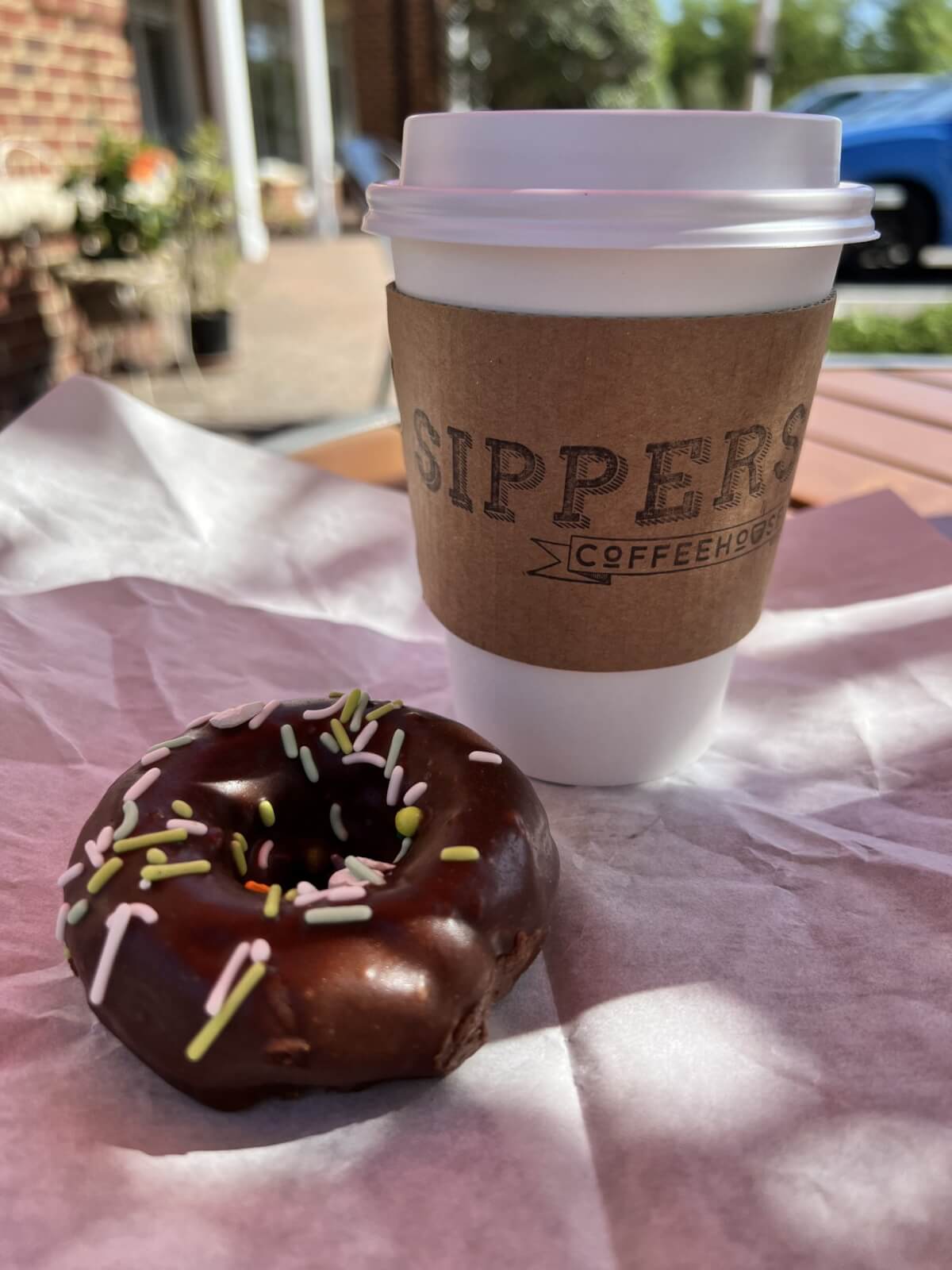 12 Florida Food & Heritage Trails Worth Traveling For - 10 A chocolate-frosted donut with sprinkles sits on parchment paper next to a takeaway coffee cup labeled "Sippers Coffeehouse"—the perfect Florida food treat to enjoy while exploring local heritage trails.