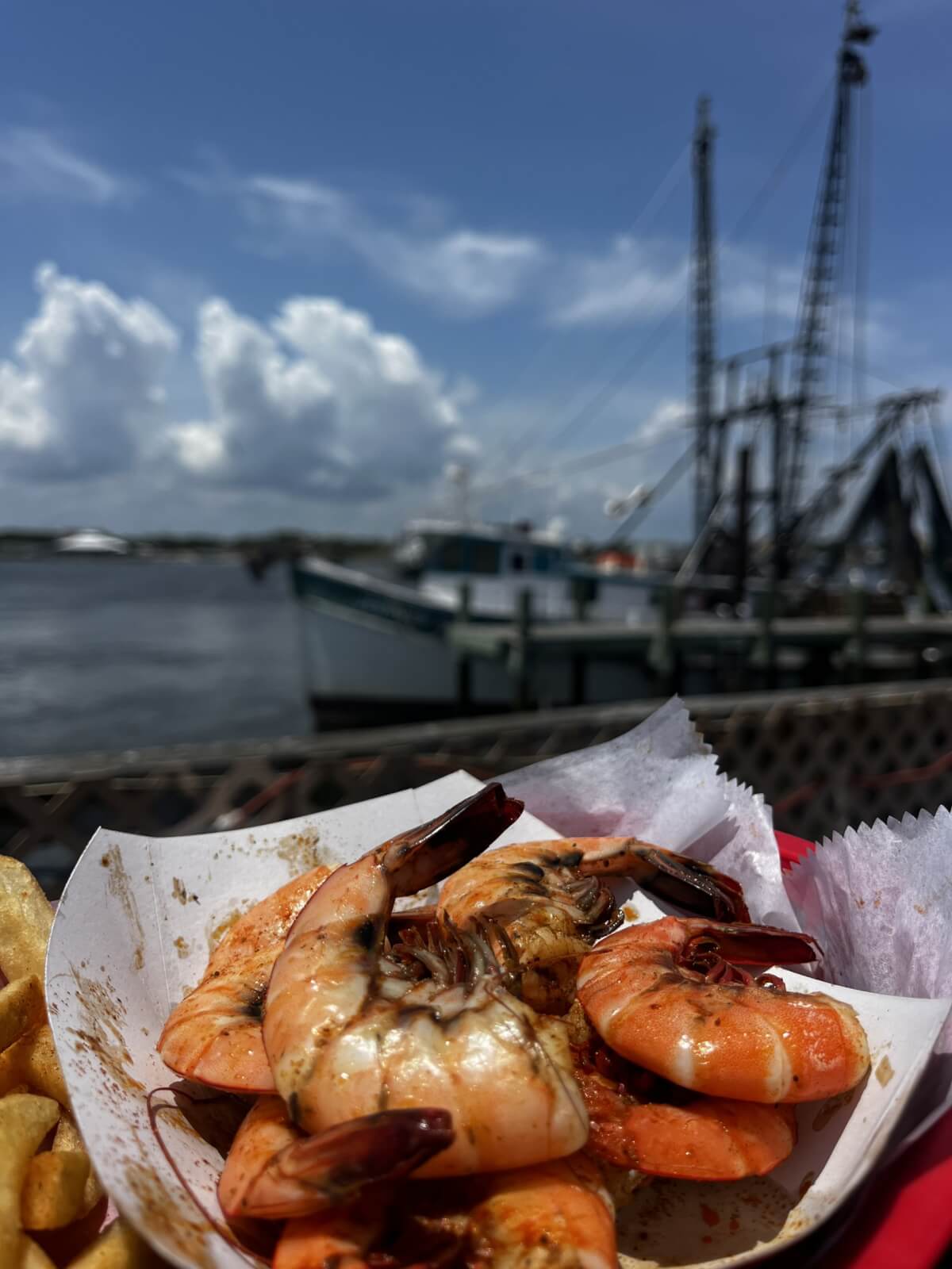 12 Florida Food & Heritage Trails Worth Traveling For - 9 A paper tray of cooked shrimp sits on a table beside potato chips, with boats and water in the background under a partly cloudy sky—a perfect scene to enjoy classic Florida food while exploring Heritage Trails.