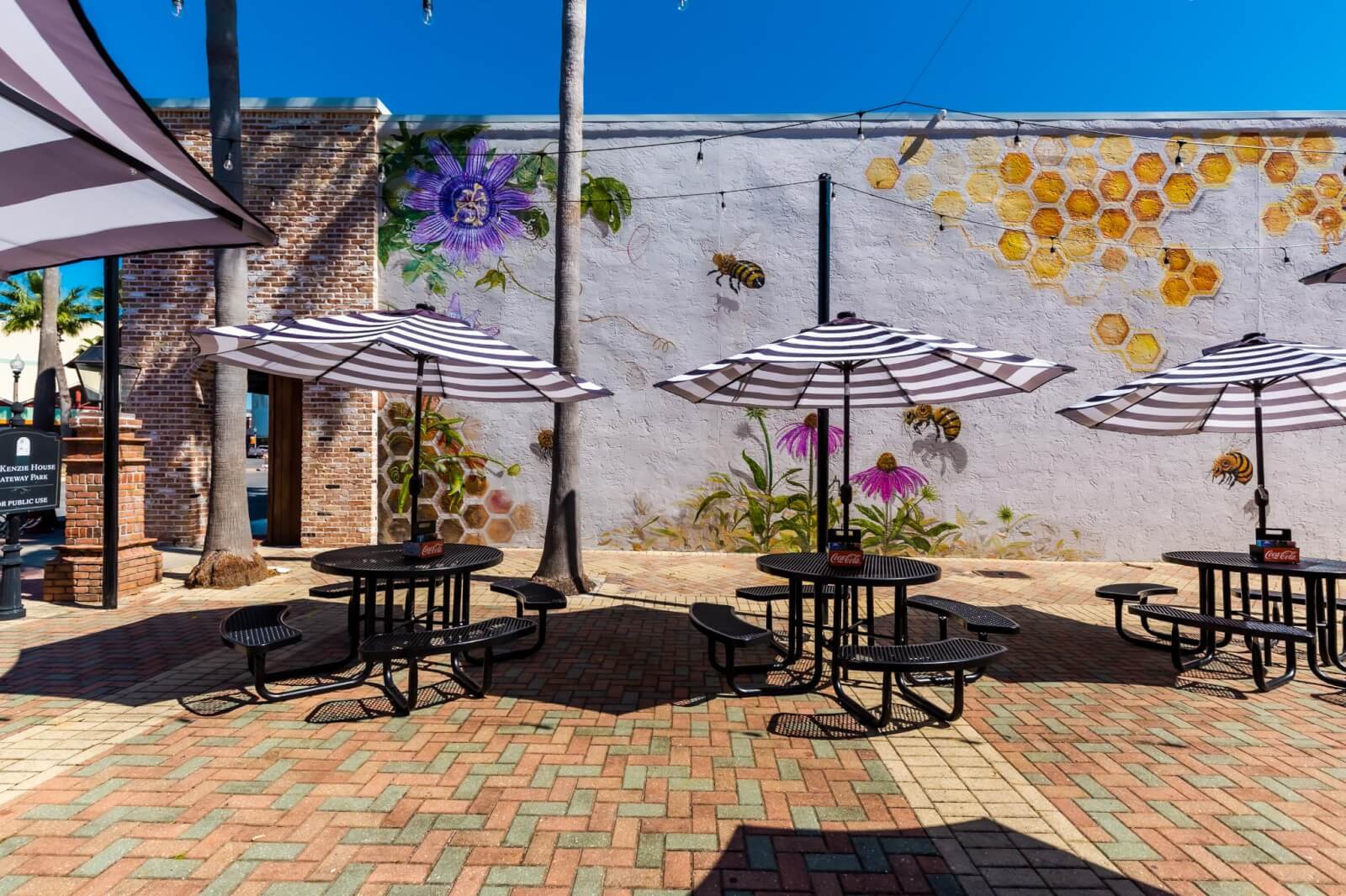 12 Florida Food & Heritage Trails Worth Traveling For - 6 Outdoor patio area with round tables and striped umbrellas on brick pavement, next to a mural of flowers, bees, and honeycombs—perfect for a relaxing Florida Food experience or a Heritage Trails stop.