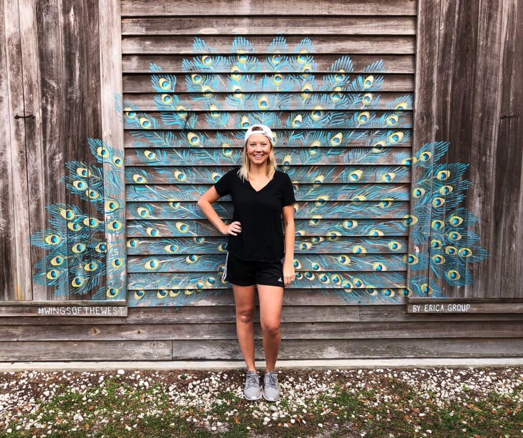 12 Florida Food & Heritage Trails Worth Traveling For - 5 A woman in black athletic wear stands in front of a wooden wall painted with a vibrant peacock feather mural, giving the illusion of peacock feathers behind her—a perfect travel snapshot along Florida’s colorful Heritage Trails.