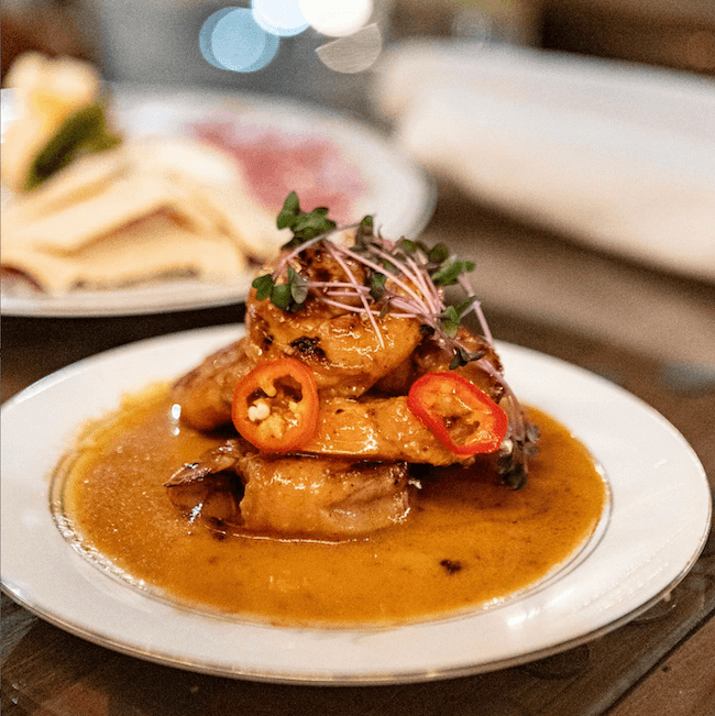 Oxford's New Must-Visit Cocktail Bar: Miscellanea Spirit House - 6 A plate of grilled shrimp in sauce, garnished with sliced red chili and microgreens, served on a white dish—perfect fare for an evening at an Oxford cocktail bar. A blurred plate of flatbread is in the background.