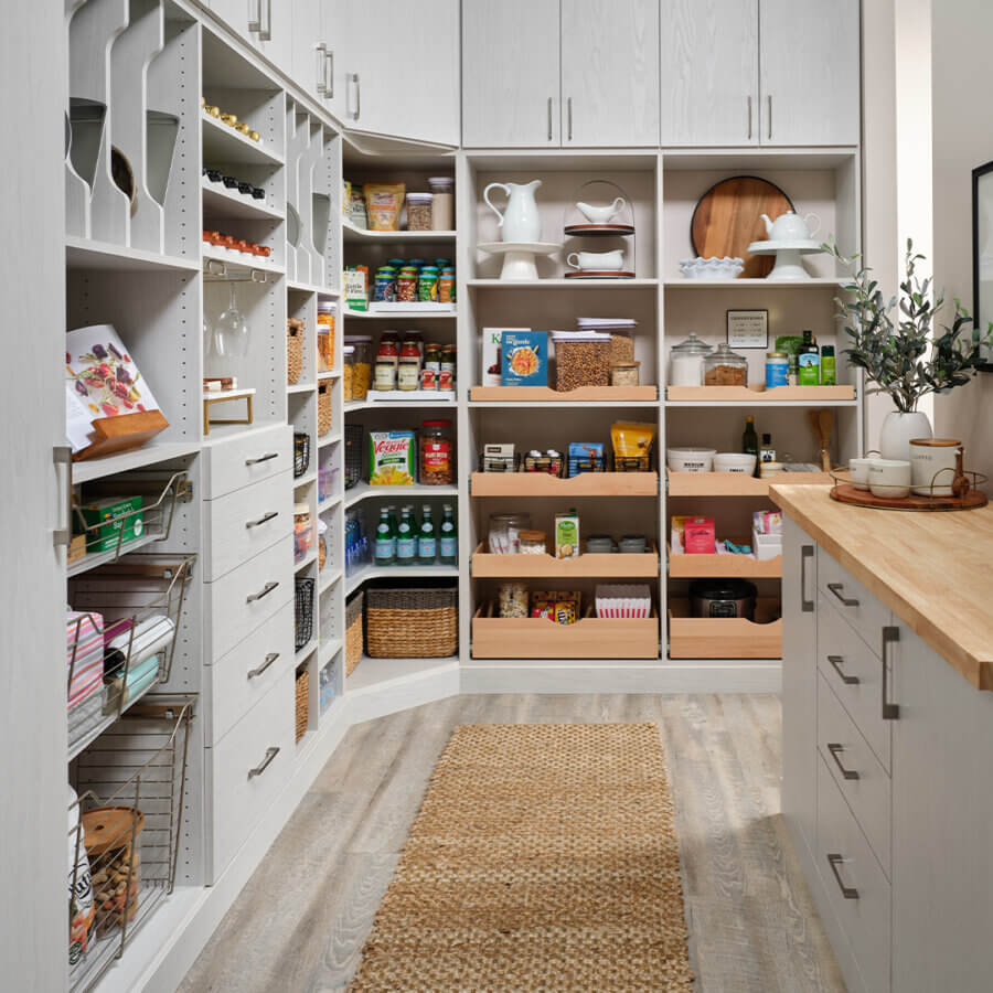 Custom Organization Ideas Homeowners Are Loving Right Now - 3 A well-organized pantry showcases smart organization ideas, with shelves, baskets, and drawers holding food items, kitchenware, canned goods, and cleaning supplies. A woven rug runs along the wood floor—perfect inspiration for homeowners.