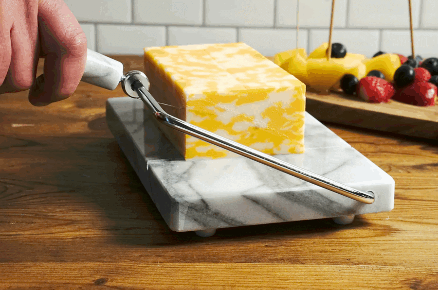 50 Under $50: Your Nashville Holiday Gift Guide - 14 A hand slices a block of marbled cheese with a wire cheese slicer on a marble board; assorted fruit is visible in the backgroundβperfect for Nashville gatherings or as a chic pick from the Holiday Gift Guide: gifts under $50.