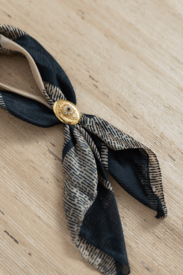 50 Under $50: Your Nashville Holiday Gift Guide - 26 A patterned black and beige scarf tied with a gold decorative pin rests on a light wooden surfaceβan elegant choice for gifts under $50 in any Holiday Gift Guide.