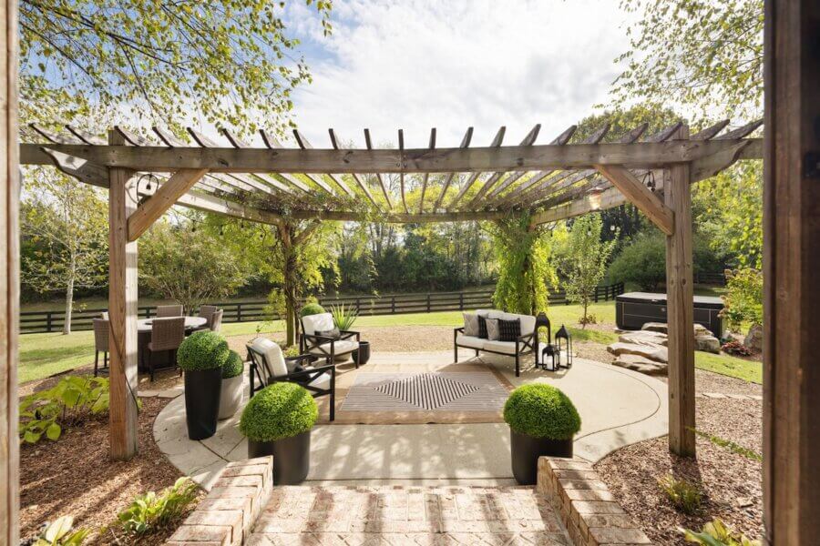 Just Listed! A Franklin Home Where Style & Serenity Coexist - 10 Wooden pergola with seating area, potted plants, and decorative lanterns on a circular patio creates pastoral peace in the landscaped backyard of this Franklin home surrounded by trees and a fence.