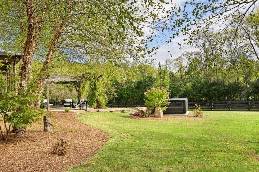 Just Listed! A Franklin Home Where Style & Serenity Coexist - 11 A landscaped backyard at this Franklin Home offers pastoral peace with a lawn, mulch path, trees, pergola seating, hot tub, and a black wooden fence—perfect for luxury living.