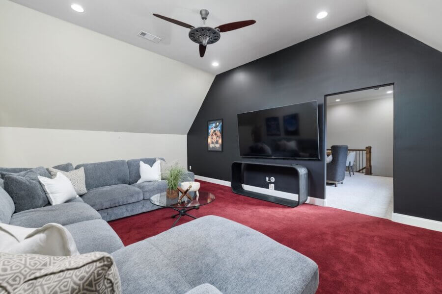 Just Listed! A Franklin Home Where Style & Serenity Coexist - 15 Experience Luxury Living in this spacious home theater room featuring a gray sectional sofa, glass coffee table, wall-mounted TV, black accent wall, and plush red carpet flooring—an inviting retreat in your Franklin Home.