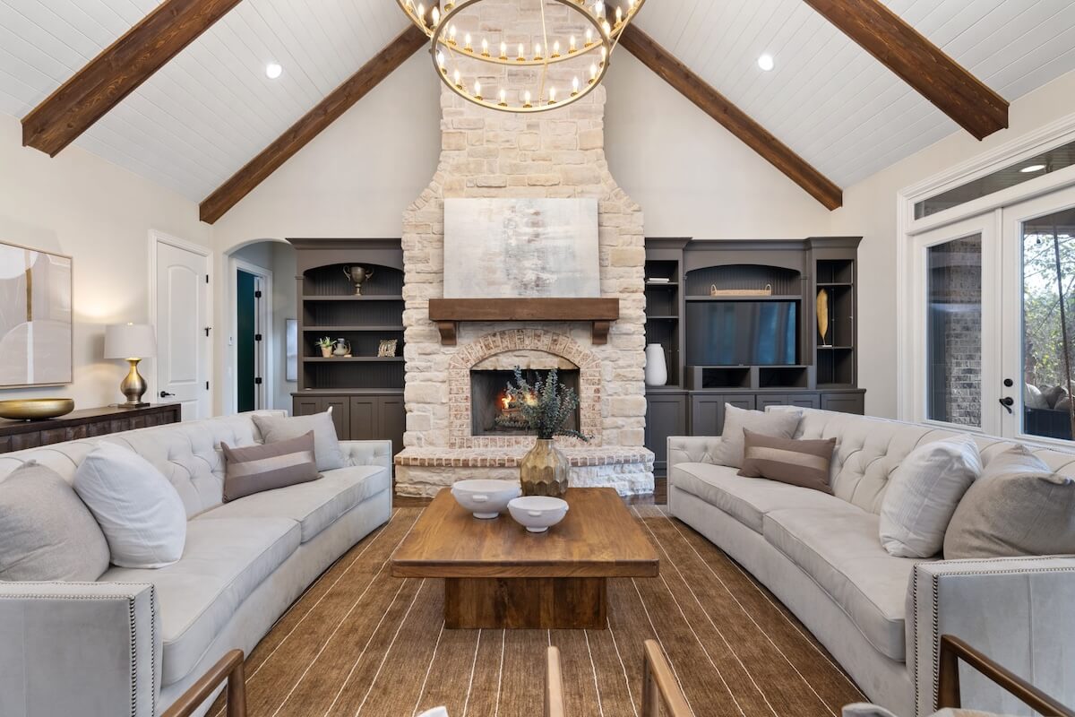 A spacious living room in this Franklin home features two gray sofas, a wooden coffee table, a stone fireplace, built-in shelves, and a large chandelier under a vaulted ceiling with exposed beams—perfect for luxury living.