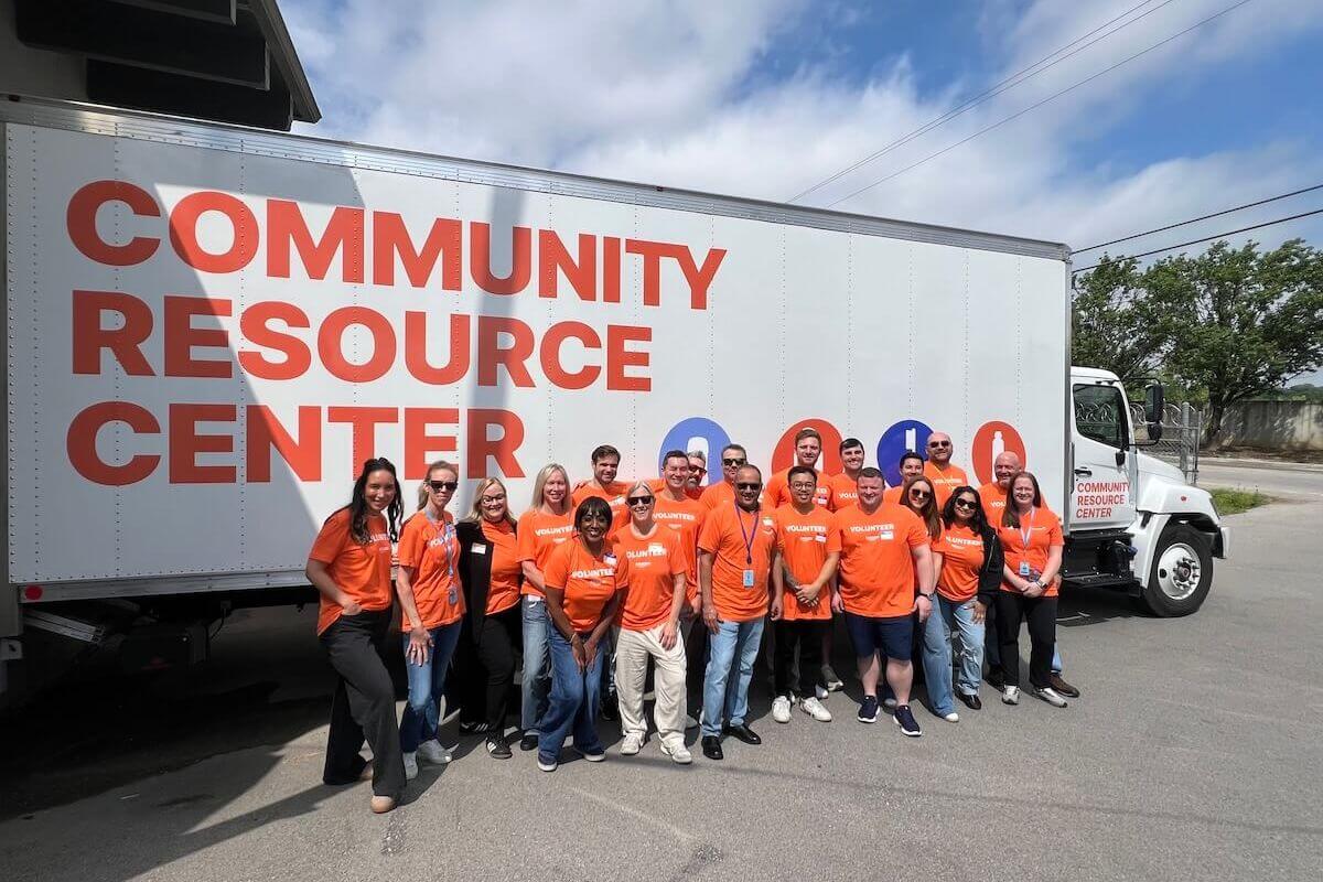 A group of people wearing orange shirts stand in front of a truck labeled "Community Resource Center," ready to lend a hand and support their neighborhood on a sunny day.