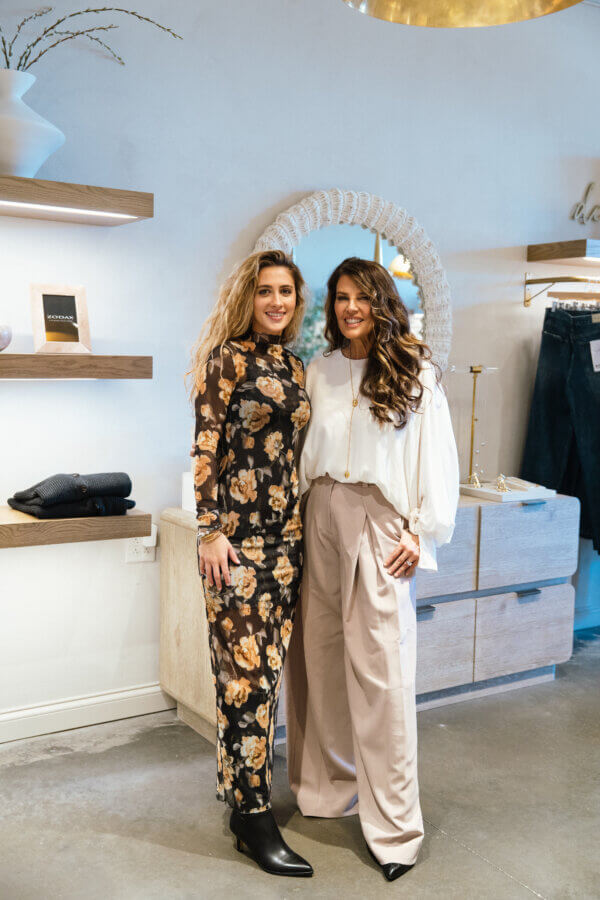 3 Franklin Boutiques Share Their Winter Wardrobe Favorites - 18 Two women stand side by side indoors, smiling in stylish winter fashion. One wears a floral dress and black boots; the other sports a white blouse with beige wide-leg pants. Shelves and decor from Franklin Boutiques are visible behind them.