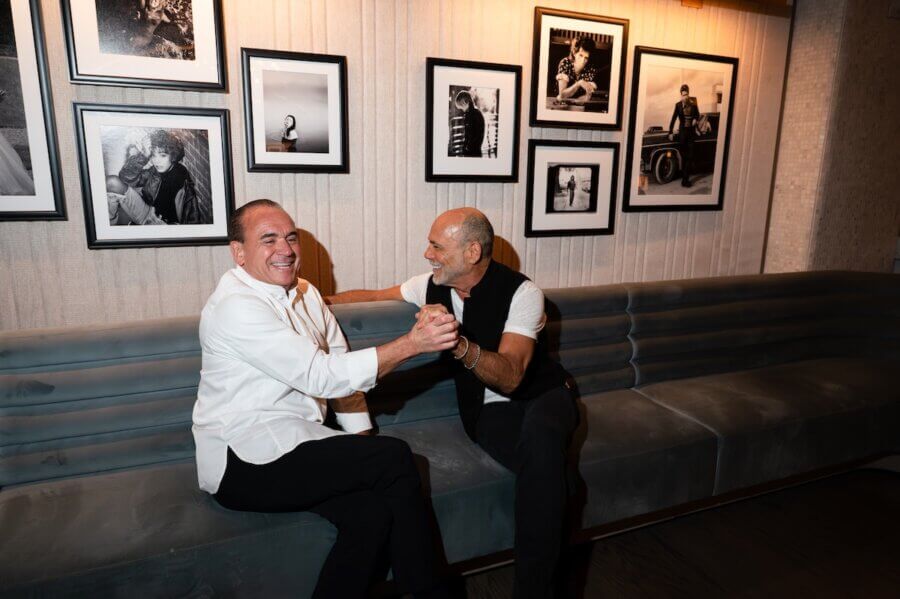 The Hermitage Hotel Debuts Exclusive Celebrity Portrait Exhibition - 5 Two men sit on a long couch at the Hermitage Hotel, smiling and clasping hands in a friendly manner, with black-and-white photos from the Exclusive Exhibition displayed on the wall behind them.