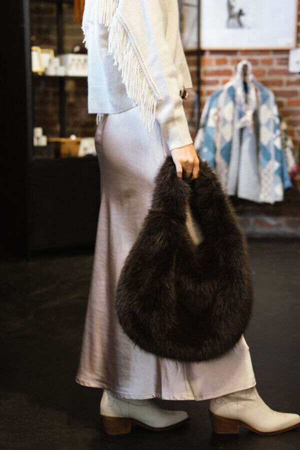 3 Franklin Boutiques Share Their Winter Wardrobe Favorites - 11 A person wearing a white fringe sweater, satin skirt, and white boots holds a large, dark furry handbag indoors—an effortless Winter Fashion look perfect for any Winter Wardrobe.