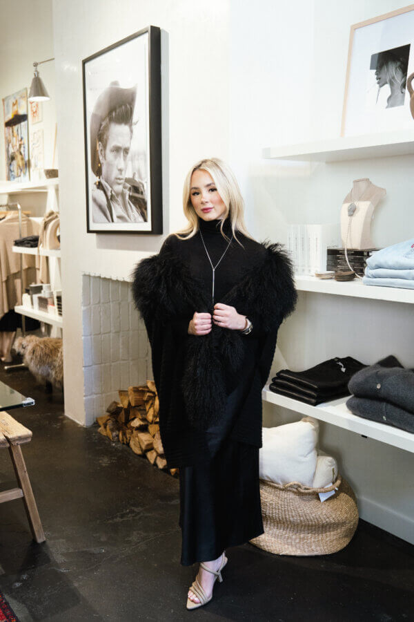 3 Franklin Boutiques Share Their Winter Wardrobe Favorites - 8 A woman in a black outfit stands in a boutique, surrounded by winter fashion essentials and accessories, with a framed photograph of a man on the wall behind her.