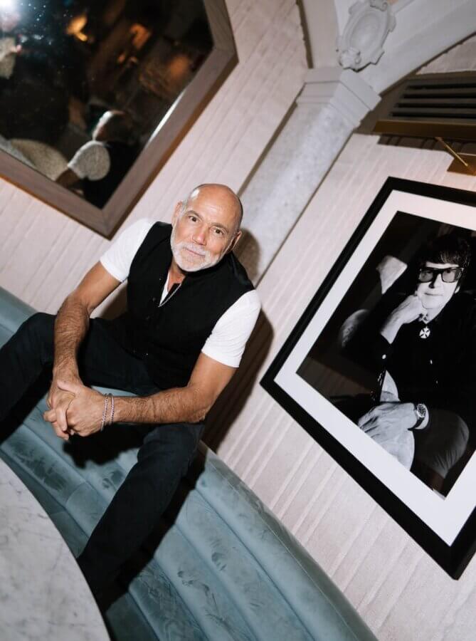 The Hermitage Hotel Debuts Exclusive Celebrity Portrait Exhibition - 2 A man with a shaved head and beard sits on a blue sofa next to a framed black-and-white portrait at the Exclusive Exhibition, held at the Hermitage Hotel.
