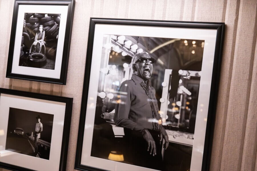 The Hermitage Hotel Debuts Exclusive Celebrity Portrait Exhibition - 4 Framed black-and-white photographs hang on a textured wall, including a central portrait of a man in sunglasses laughing in front of illuminated mirrors—part of an exclusive exhibition at the Hermitage Hotel.