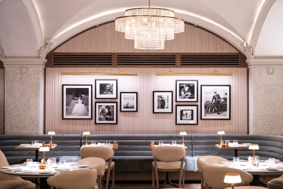 Elegant restaurant interior at the Hermitage Hotel features round tables, beige chairs, a blue cushioned bench, small table lamps, and framed black-and-white photos from an exclusive exhibition under a large chandelier.