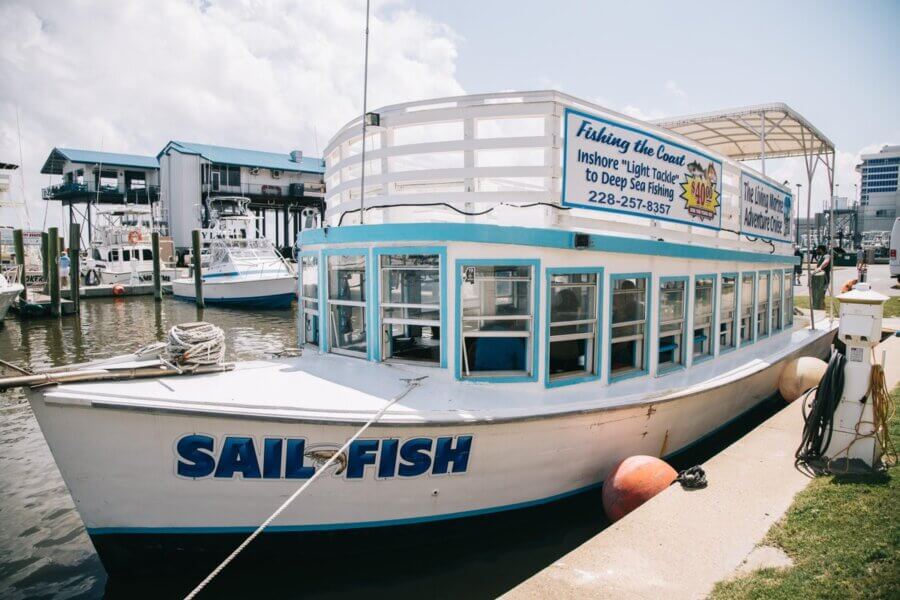 A white and blue boat named "Sail Fish" is docked at a marina, the perfect starting point for a Culinary Adventure along the coast, with buildings and other boats visible in the background.