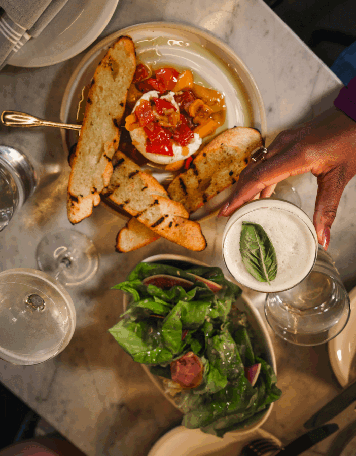 A hand holds a cocktail garnished with basil beside grilled bread topped with cheese and tomatoes, and a spinach salad on a marble table—a true Mississippi Eats culinary adventure.