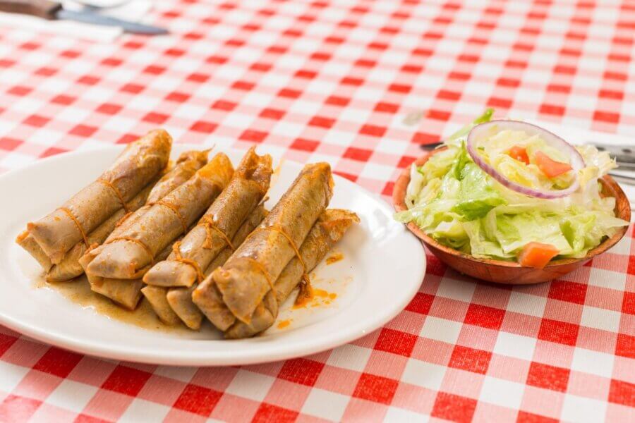 A plate with five rolled taquitos topped with sauce and a side bowl of lettuce salad with tomato and onion on a red and white checkered tablecloth—perfect for a Bucket List Mississippi Eats culinary adventure.