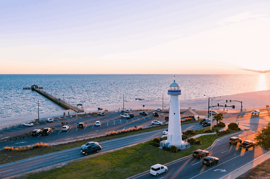 A white lighthouse stands near a road by the beach, with cars driving and parked; a pier extends into the calm ocean at sunset—a classic scene of Southern Tradition perfect for your Bucket List Mississippi Eats culinary adventure.