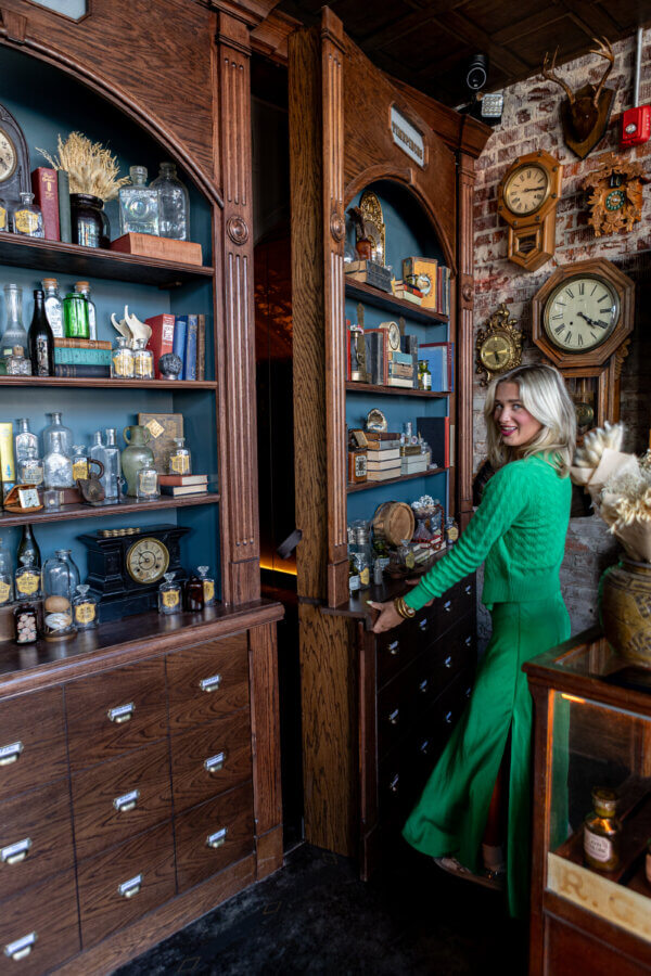 Oxford's New Must-Visit Cocktail Bar: Miscellanea Spirit House - 3 A woman in a green outfit opens a hidden door disguised as a bookshelf in a vintage room at Miscellanea, an Oxford cocktail bar filled with clocks, bottles, and wooden cabinets—a must-visit for lovers of mystery and style.