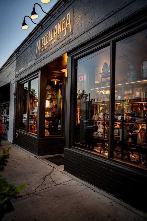 Oxford's New Must-Visit Cocktail Bar: Miscellanea Spirit House - 2 A must-visit Oxford cocktail bar, Miscellanea Spirit House features large windows showcasing shelves of bottles and decor items, all illuminated by warm indoor lighting.