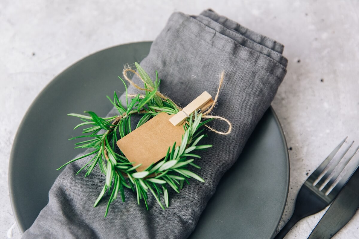 A gray plate with a gray cloth napkin, garnished with a small rosemary wreath and a blank brown paper tag clipped on, next to a black fork and knife—perfect for effortless holiday hacks or festive holiday ideas.