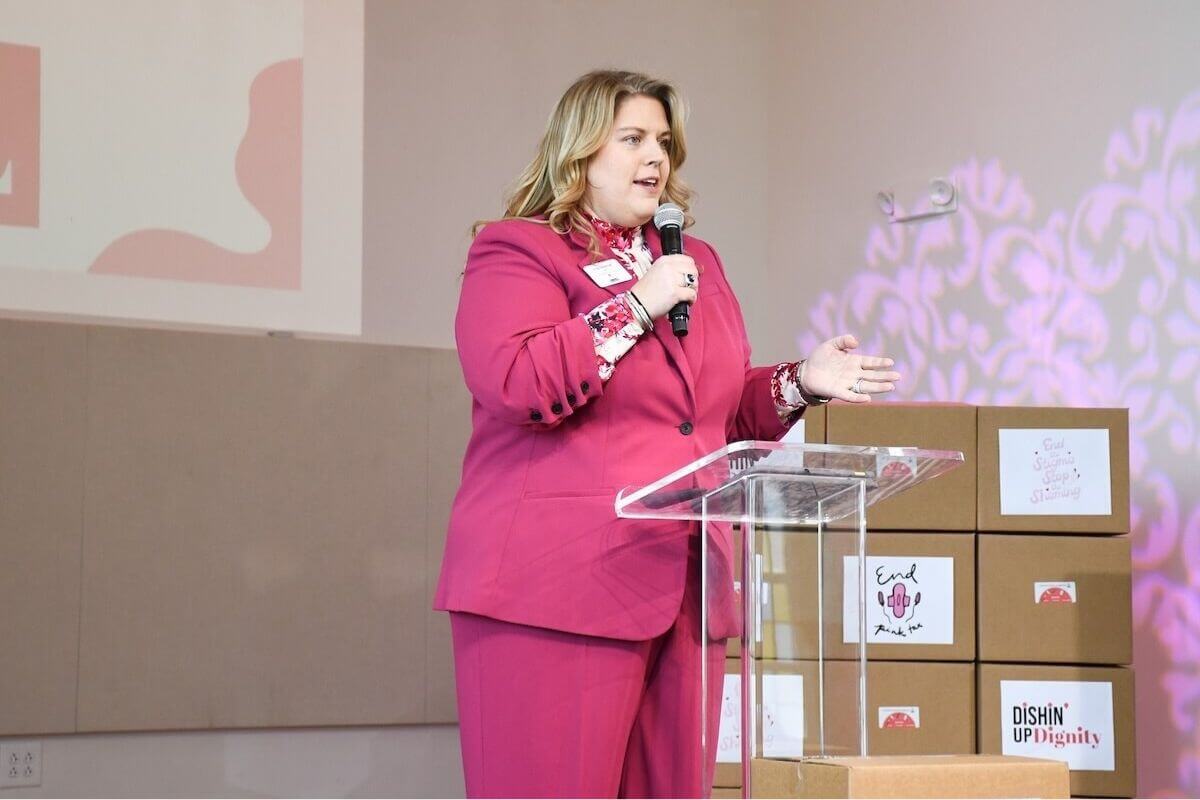 A woman in a pink suit speaks at a clear podium, with stacked boxes labeled "End" and "DISHIN’ UP Dignity" visible in the background at a community resource center.