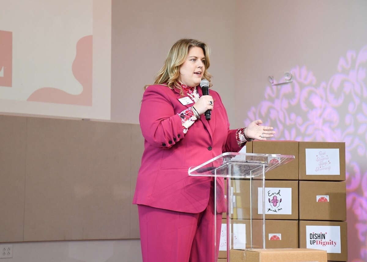 Meet the CEO of Nashville's Community Resource Center - 5 CEO Tina Doniger, in a bright pink suit, speaks at a clear podium. Stacked cardboard boxes and a projected Community Resource Center presentation are visible in the background.
