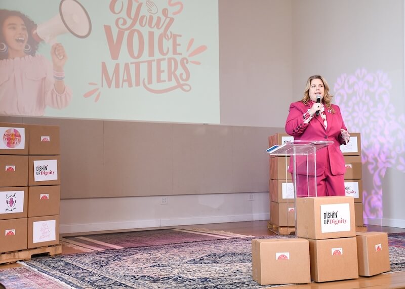 Meet the CEO of Nashville's Community Resource Center - 2 CEO Tina Doniger, in a pink suit, speaks at a podium beside stacked boxes in a room at the Community Resource Center, with a large "Your Voice Matters" sign projected on the wall behind her.