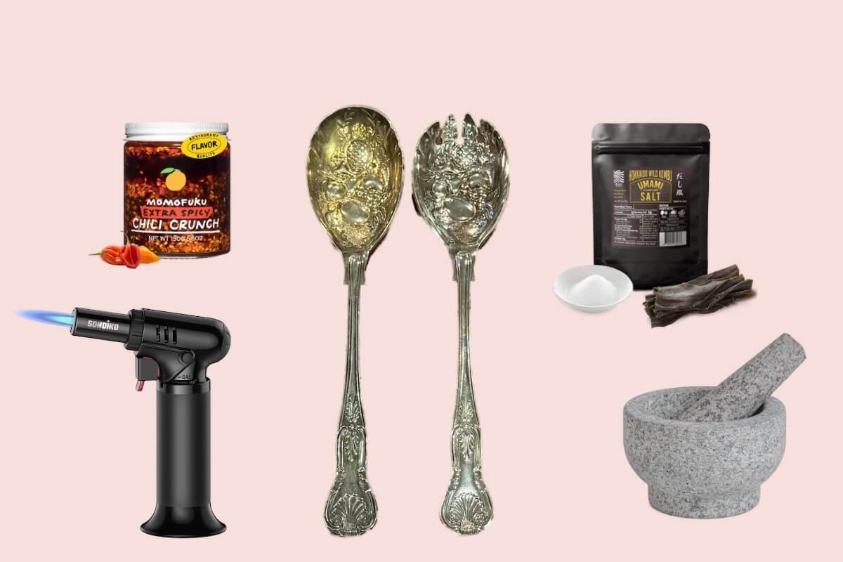 A selection of culinary gifts—including a torch, jar of chili crunch, ornate serving spoons, salt, dried seaweed, and a stone mortar and pestle—are artfully arranged on a light pink background.