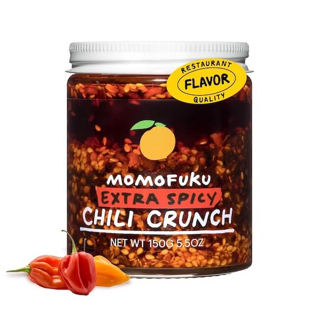 12 Nashville Chefs Share Their Go-To Culinary Gifts to Give - 7 A jar of Chef-approved Momofuku Extra Spicy Chili Crunch with two red chili peppers placed beside it on a white background—perfect for Nashville culinary fans or spicy Holiday gifts.