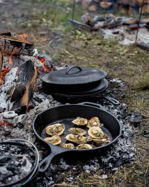 12 Nashville Chefs Share Their Go-To Culinary Gifts to Give - 2 A cast iron skillet with oysters cooking over a campfire—culinary insiders in Nashville will love this scene, with a lidded pot and pan nearby on the ground, surrounded by ashes and grass.