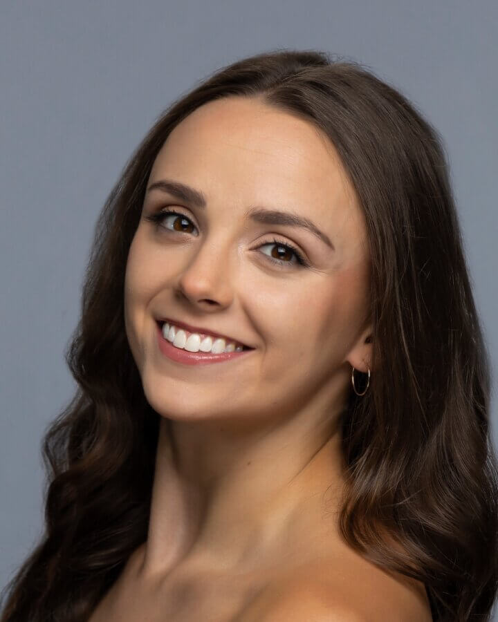 Meet the Star of Nashville Ballet’s Nutcracker - 2 A young woman with long brown hair smiles at the camera against a plain gray background. She is wearing a small hoop earring.