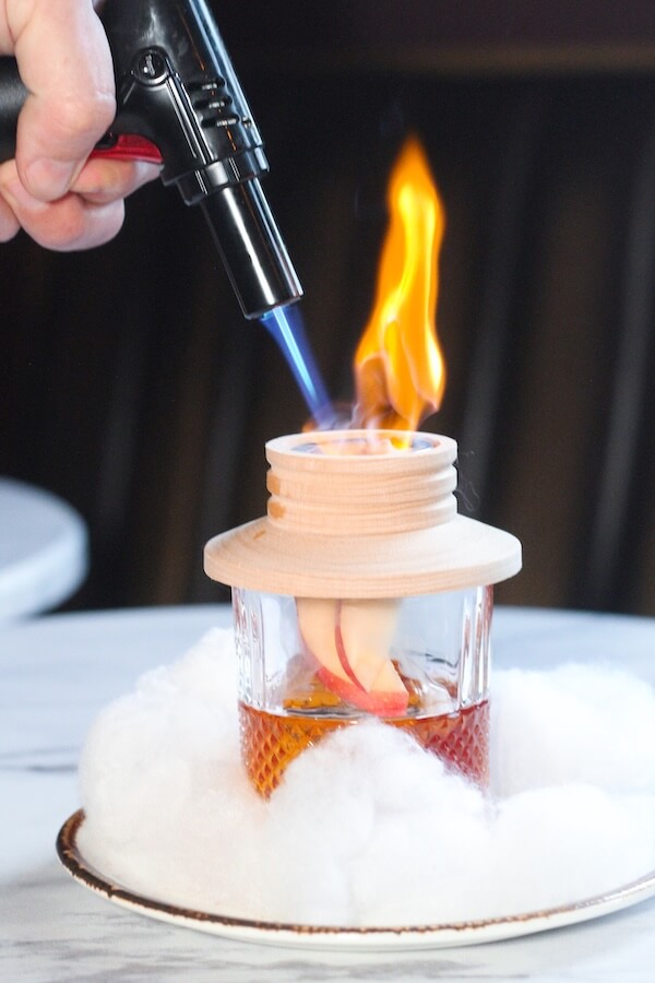 30+ Nashville Holiday Pop-Ups to Get You in the Spirit! - 5 A hand uses a blowtorch to ignite a flame above a cocktail in a glass, surrounded by white cotton-like materialβa festive touch perfect for Nashville Holiday Pop-Ups and memorable holiday events.