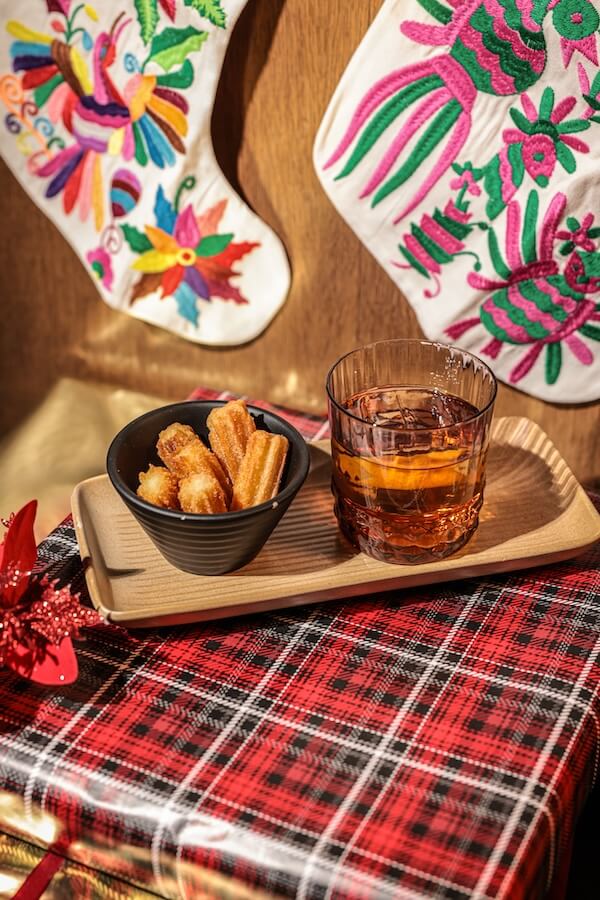 30+ Nashville Holiday Pop-Ups to Get You in the Spirit! - 10 A tray with a glass of amber-colored beverage and a bowl of churros sits on a red plaid surface, capturing the cozy spirit of Nashville Holiday Pop-Ups, with embroidered stockings hanging in the background.