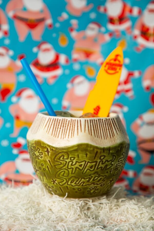 30+ Nashville Holiday Pop-Ups to Get You in the Spirit! - 12 A green ceramic cup shaped like a coconut with βSippinβ Santaβ written on it, filled with a drink, blue straw, yellow surfboard stirrer, and a festive Santa-themed backgroundβperfect for Nashville Holiday Pop-Ups and festive Nashville Christmas events.