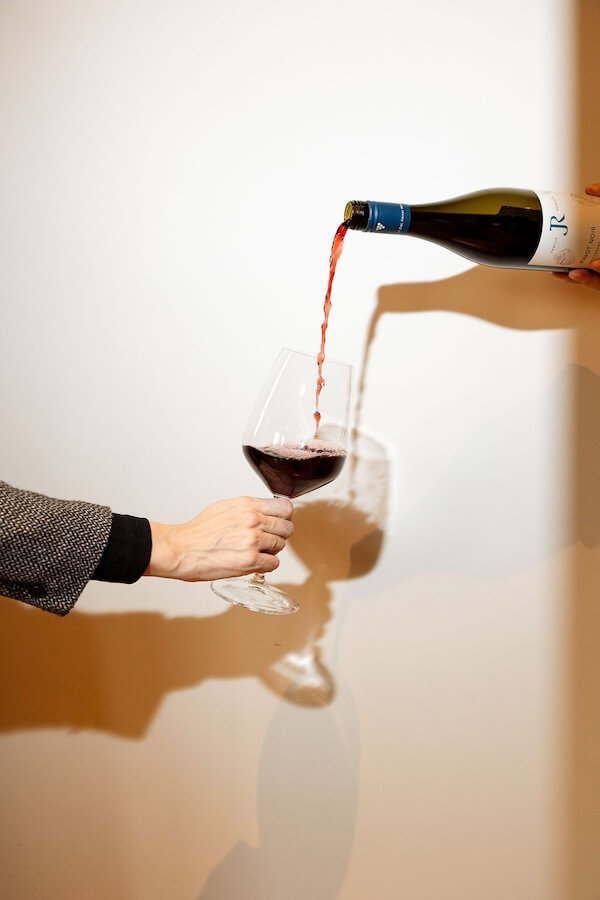 Merlot is Back?! What Nashville Will Be Drinking in 2026, According to an Expert - 4 A person pours red wine from a bottle into a wine glass held by another against a white wall, reflecting the evolving drinking trends expected in Nashville by 2026.