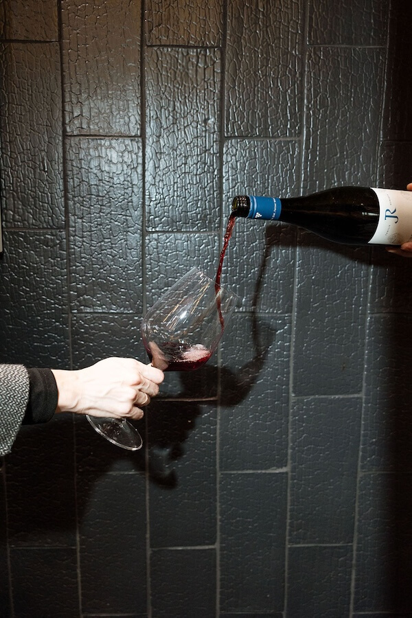 Merlot is Back?! What Nashville Will Be Drinking in 2026, According to an Expert - 2 A person pours red wine from a bottle into a wine glass against a textured dark tiled wall, reflecting Nashville's 2026 drinking trends.