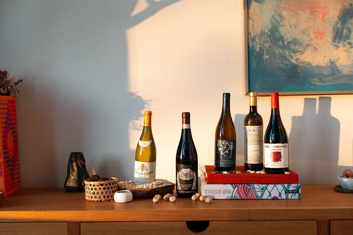 Five assorted wine bottles are displayed on a wooden sideboard with books, a bowl, nuts, and decorative items—capturing Nashville's latest drinking trends for 2026—set against a light-colored wall with a painting.