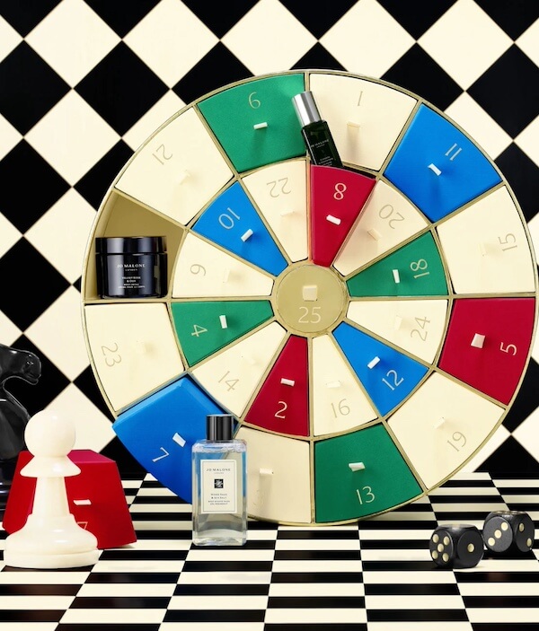 The BEST Advent Calendars for 2025 - 6 A circular advent calendar with numbered doors stands amid chess pieces, a perfume bottle, and a jar, all arranged on a black-and-white checkered background—a unique take on traditional advent calendars.