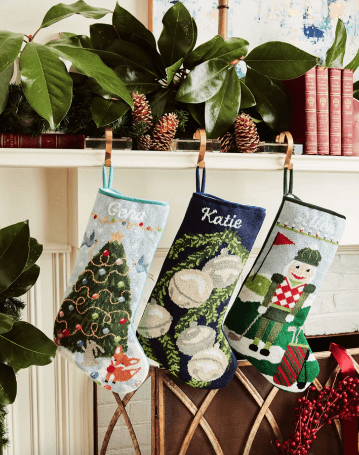 14 Southern Ornament & Stocking Shops We Love - 3 Three embroidered Christmas stockings, adorned with southern ornaments, hang from a decorated mantel with pinecones, leaves, and books; stockings feature a tree, magnolia flowers, and a nutcracker.