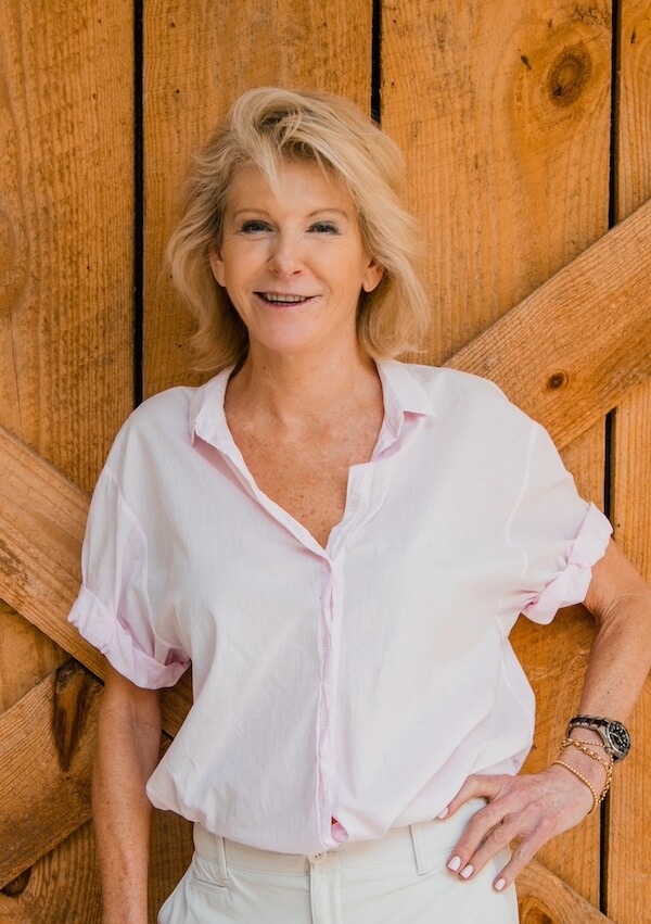 Meet Kim Wilson of Atlanta's Beloved Lucy's Market - 2 A woman with blonde hair wearing a light pink button-up shirt stands in front of a wooden wall at Lucy's Market, smiling with one hand on her hip.