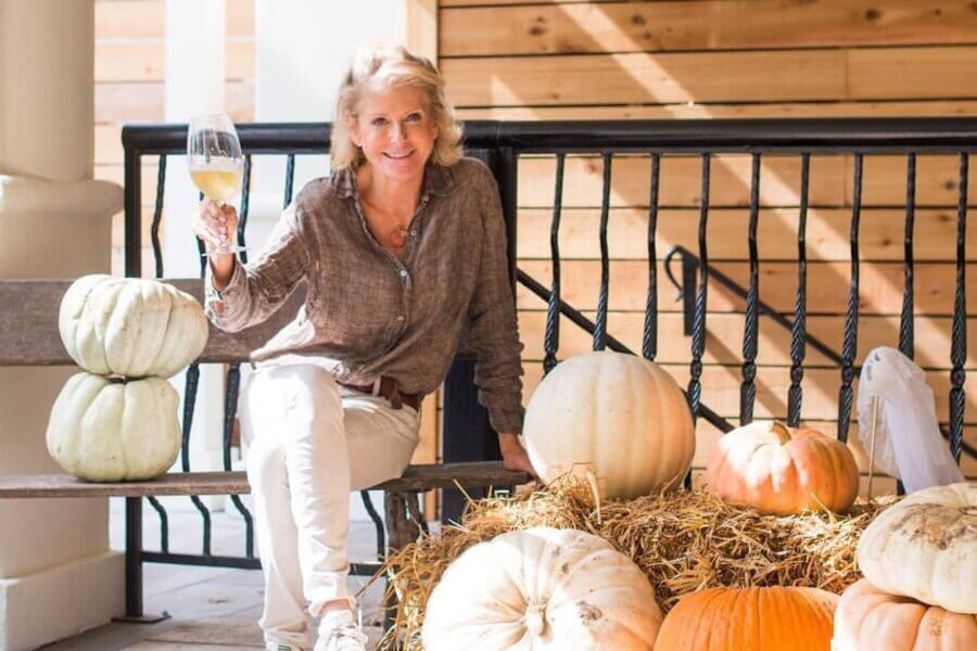 Meet Kim Wilson of Atlanta's Beloved Lucy's Market - 5 Kim Wilson sits on a bench next to a stack of pumpkins and hay at Lucy's Market, holding up a glass of white wine, with a wooden wall and railing in the background.
