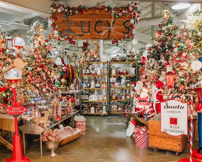 Meet Kim Wilson of Atlanta's Beloved Lucy's Market - 4 Christmas-themed store display at Lucy's Market with decorated trees, gifts, ornaments, and a sign reading "Santa comes in 53 days," surrounded by festive decorations and twinkling lights.