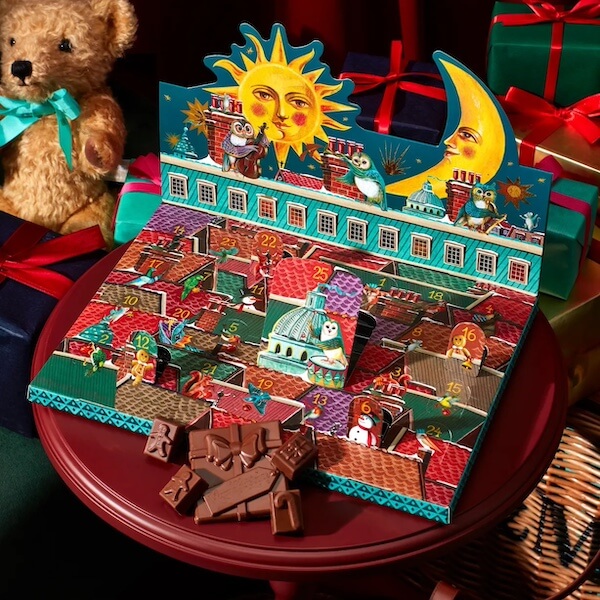 The BEST Advent Calendars for 2025 - 13 A 2025 Advent Calendar with numbered doors and whimsical rooftop illustrations sits on a round table, chocolate pieces in front, with wrapped gifts and a teddy bear in the background.
