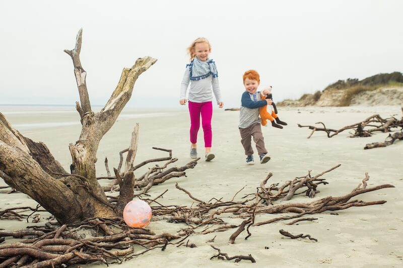 Can't-Miss 2026 Events in Georgia's Beloved Golden Isles - 2 Two children walk on a sandy beach near driftwood on the Golden Isles; the girl wears pink leggings and the boy holds a stuffed animal. A ball rests among the driftwood, capturing the spirit of travel in 2026.