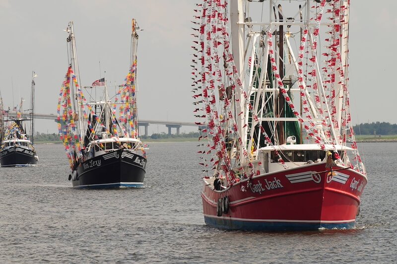 Can't-Miss 2026 Events in Georgia's Beloved Golden Isles - 8 A fleet of decorated fishing boats sails on a river, with colorful flags draped over the vessels and a bridge visible in the background—capturing the vibrant spirit of Golden Isles events.