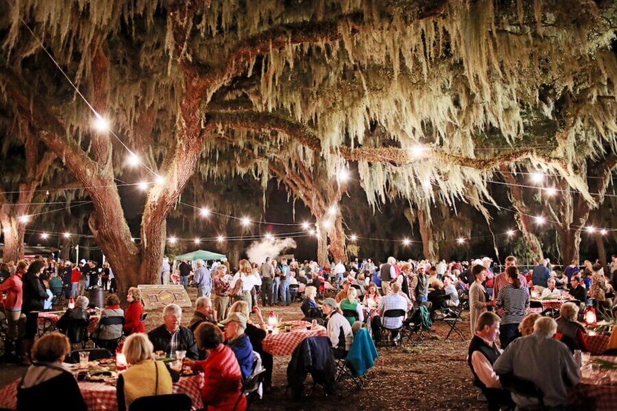 Can't-Miss 2026 Events in Georgia's Beloved Golden Isles - 4 Large group of people dining at picnic tables under trees with hanging moss and string lights at an outdoor evening event—perfect for those seeking memorable Golden Isles events and travel experiences in 2026.